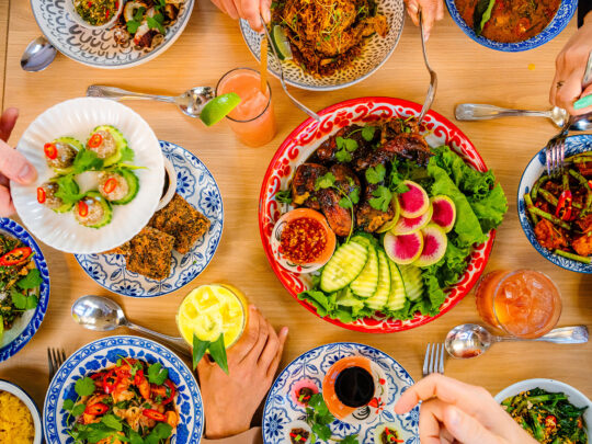 A vibrant spread of colorful Thai dishes, including plates of fresh vegetables, grilled meats, curries and dumplings, is beautifully arranged on a wooden table at Kalaya.