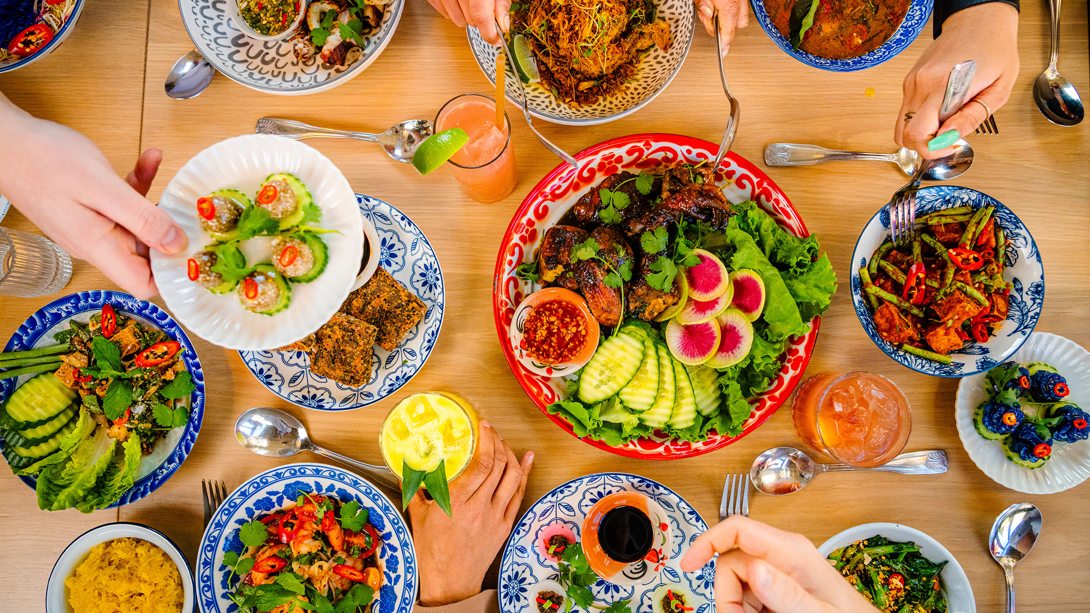 A vibrant spread of colorful Thai dishes, including plates of fresh vegetables, grilled meats, curries and dumplings, is beautifully arranged on a wooden table at Kalaya.