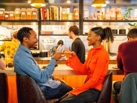 A man wearing a blue top and a woman wearing a vibrant orange blouse clink glasses while seated at the bar at Laser Wolf.