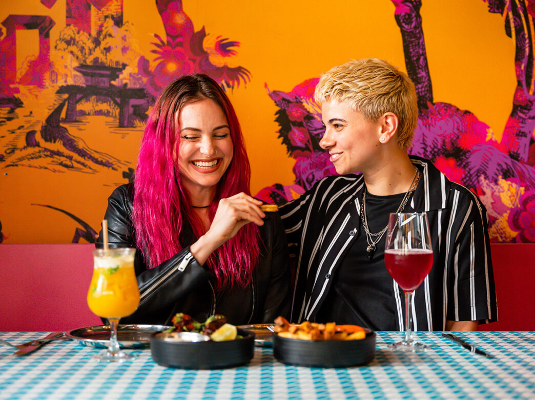 Two people, seated in front of a vibrant pink and orange mural, share a laugh over cocktails and small plates at Laser Wolf.