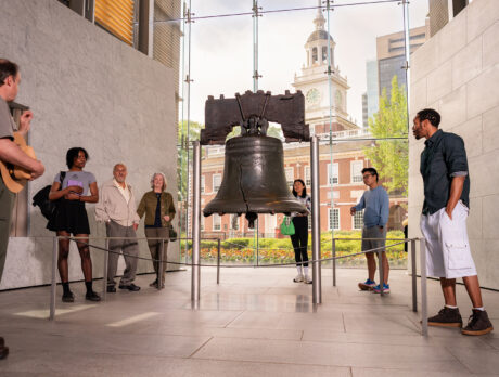 Seven people, including a National Park Service ranger, stand around and observe the Liberty Bell at Liberty Bell Center. Independence Hall is seen through the floor to ceiling windows behind the Liberty Bell.