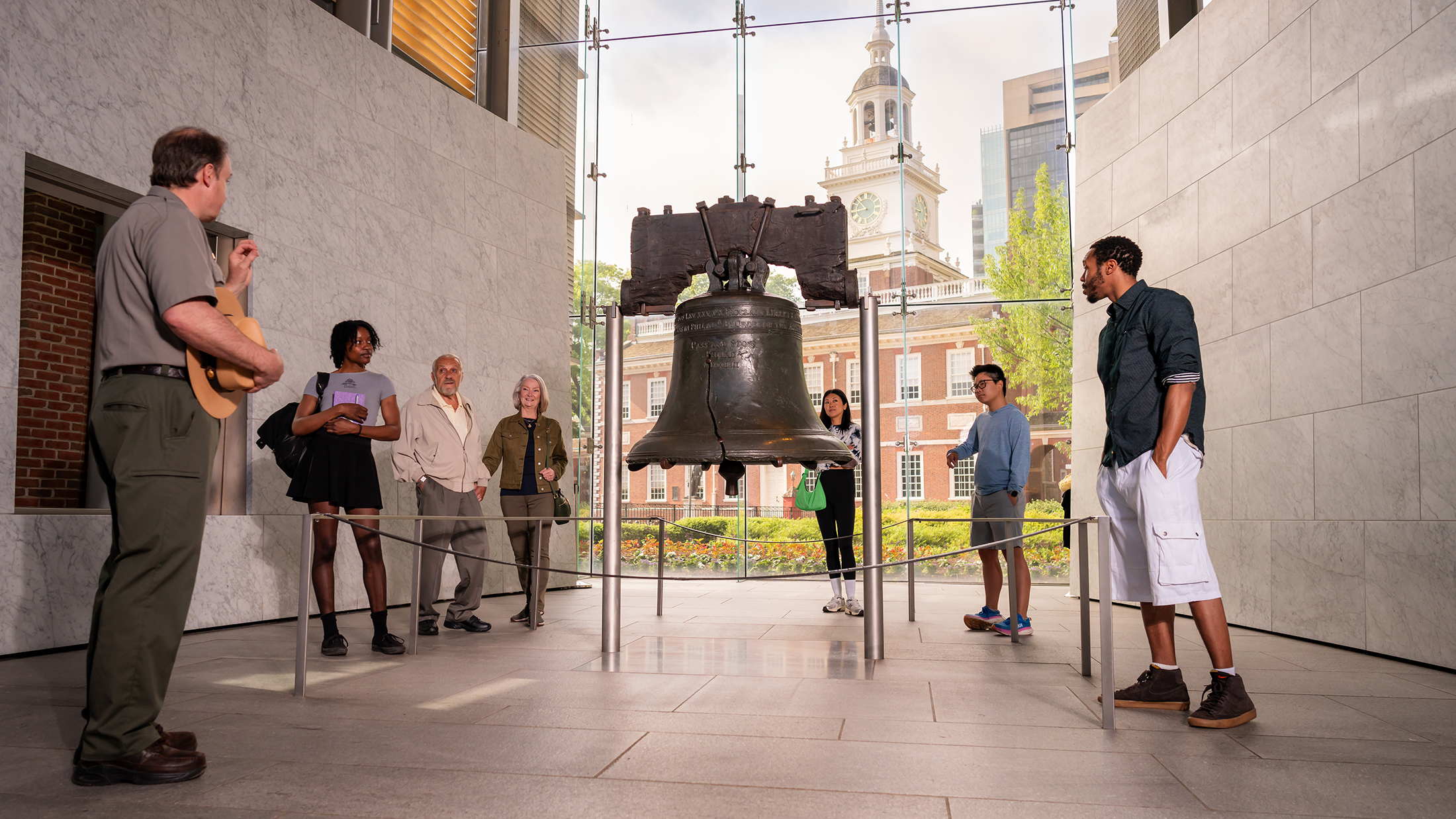 Seven people, including a National Park Service ranger, stand around and observe the Liberty Bell at Liberty Bell Center. Independence Hall is seen through the floor to ceiling windows behind the Liberty Bell.