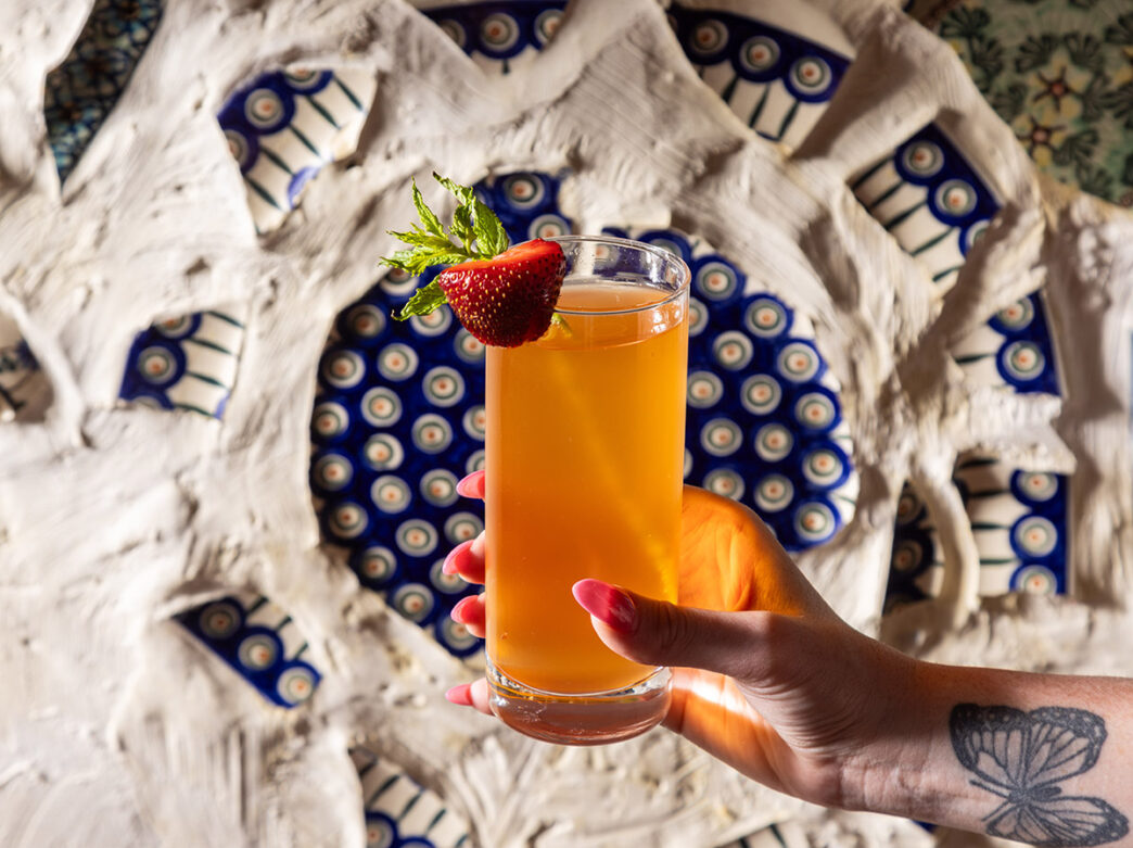 A vibrant orange cocktail garnished with a fresh strawberry is held against a textured wall adorned with intricate blue and white ceramic accents at Little Walter’s.