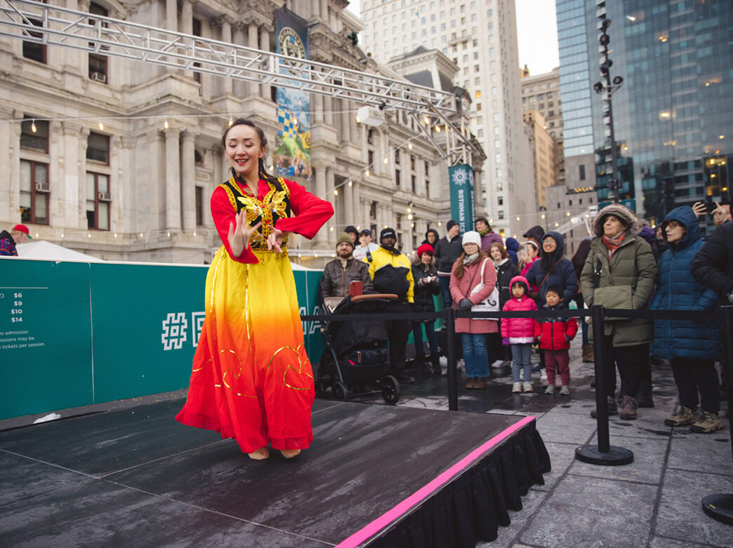 A traditional Chinese dancer performs on a black stage in Dilworth Park. City Hall is in the background and people gather around the stage to watch.