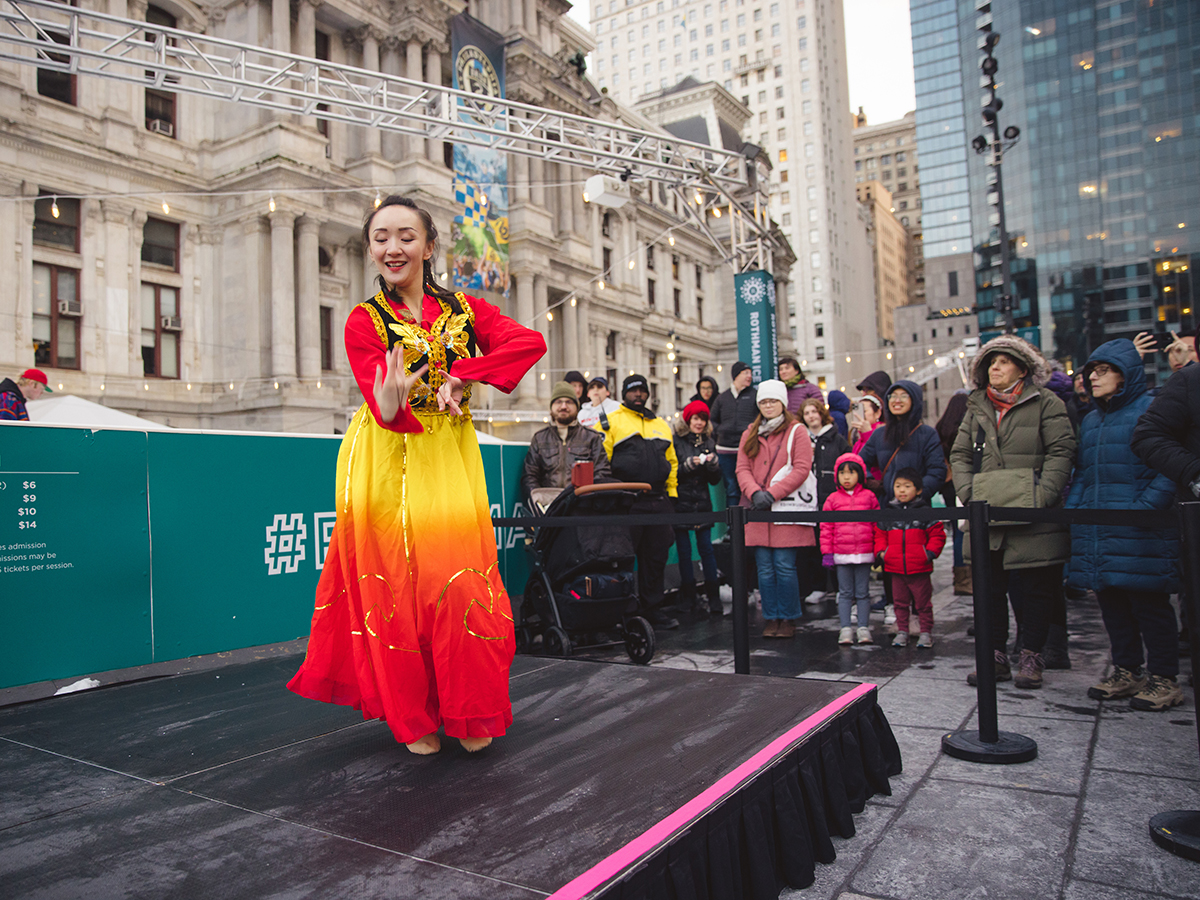 A traditional Chinese dancer performs on a black stage in Dilworth Park. City Hall is in the background and people gather around the stage to watch.