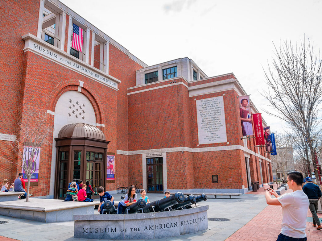 A grande fachada de tijolos do Museum of the American Revolution se destaca enquanto os visitantes aproveitam os assentos e os canhões decorativos na praça externa.