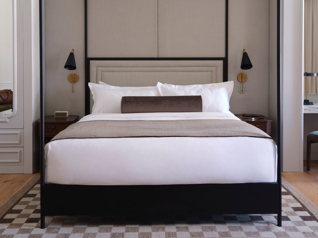 A black metal four poster bed sits in the middle of a one bedroom suite at The Bellevue Hotel. The bed has white linens and a brown lumbar pillow.