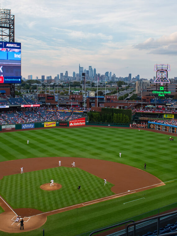 A panoramic view of the Phillies game at Citizens Bank Park showcases the vibrant green field, the towering scoreboard, and the Philadelphia skyline in the distance.