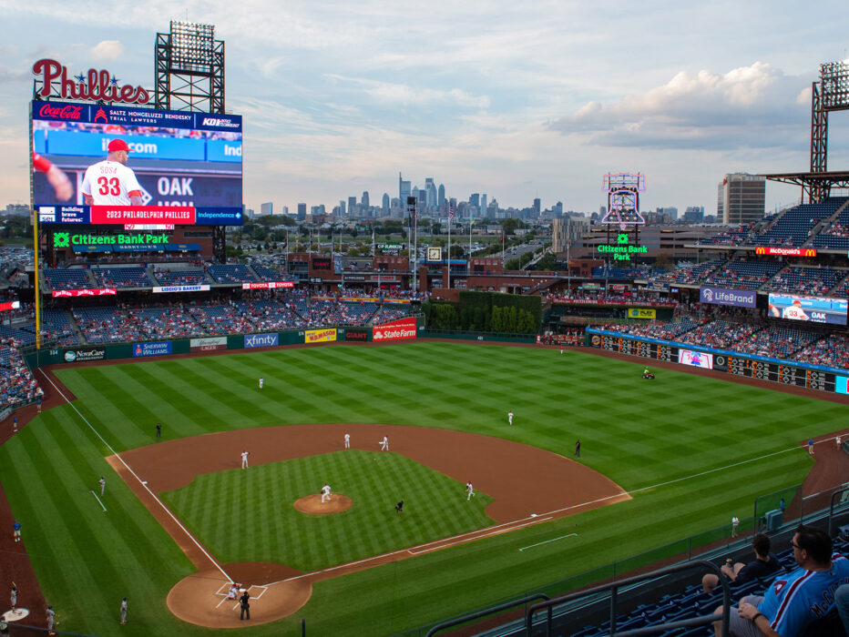 A panoramic view of the Phillies game at Citizens Bank Park showcases the vibrant green field, the towering scoreboard, and the Philadelphia skyline in the distance.