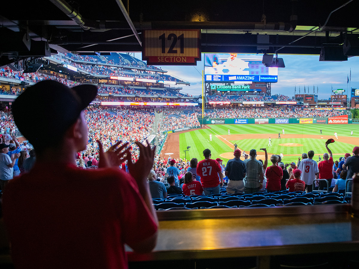 A young fan watching the game from a shaded section at Citizens Bank Park claps enthusiastically, joining the lively crowd as they cheer on the Phillies.