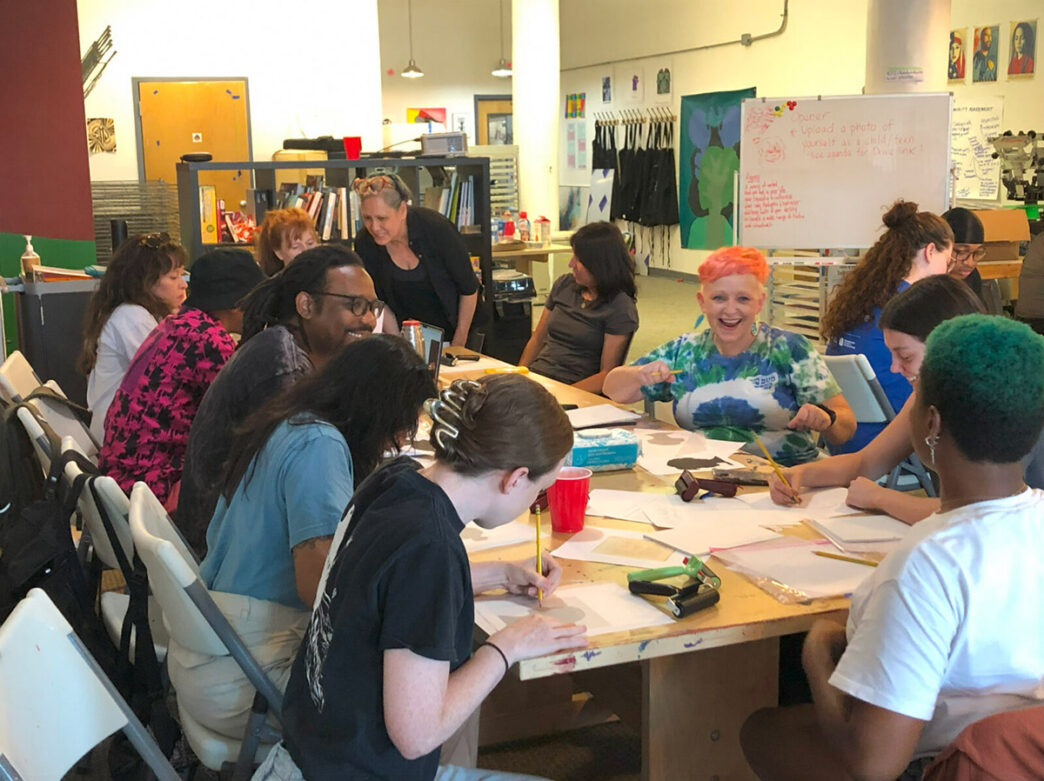 Smiling participants gather around a table at a Mural Arts workshop, actively engaged in printmaking and creative discussions.