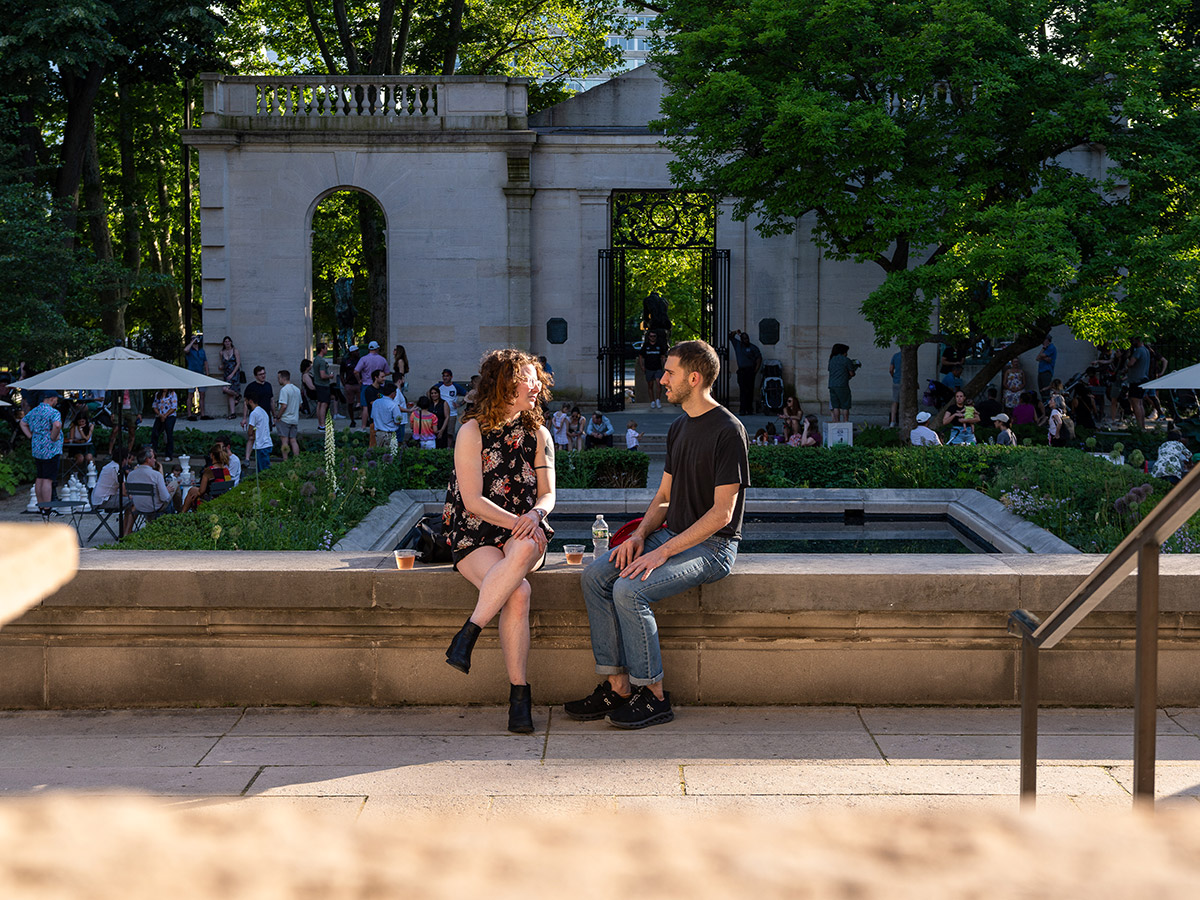 A couple sits on the stone ledge of the Rodin Museum garden, enjoying a casual moment with drinks in hand. Behind them, the museum's elegant stone archway and lush greenery frame a lively scene of people mingling and relaxing around tables and a reflecting pool.