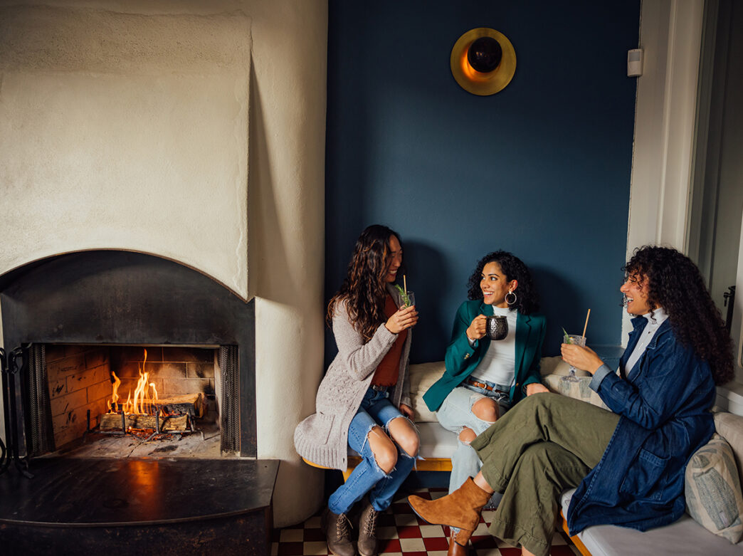 Three women smile and enjoy drinks while sitting on a bench next to a cozy fireplace in Suraya.
