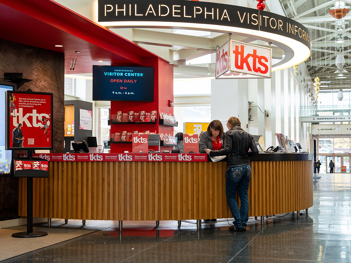 The new TKTS counter inside the Independence Visitor Center, featuring a modern red and wood design. A staff member assists a visitor at the counter, surrounded by signage and brochures promoting discounted show tickets.