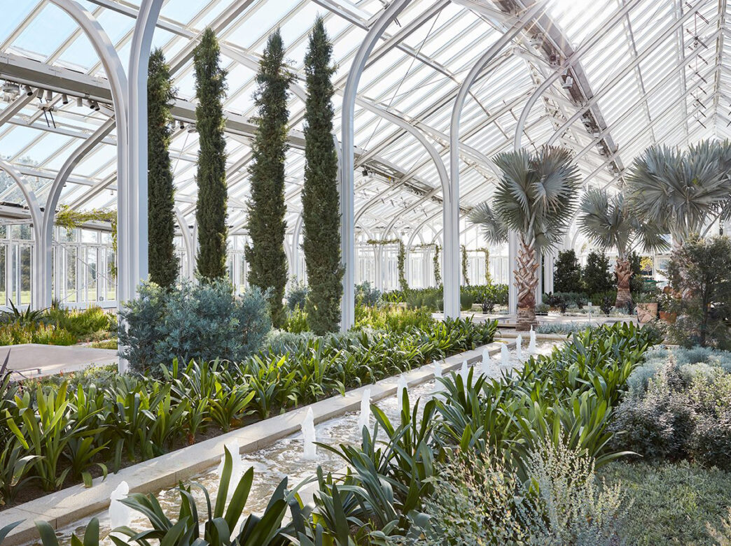 An interior view of the new West Conservatory at Longwood Gardens. Various plants, palm trees and water features fill the glass greenhouse.