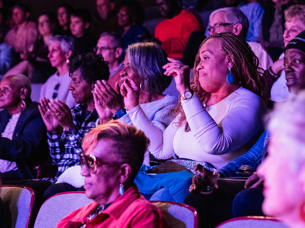 People seat in the audience and clap their hands while watching a production at Theatre Horizon.
