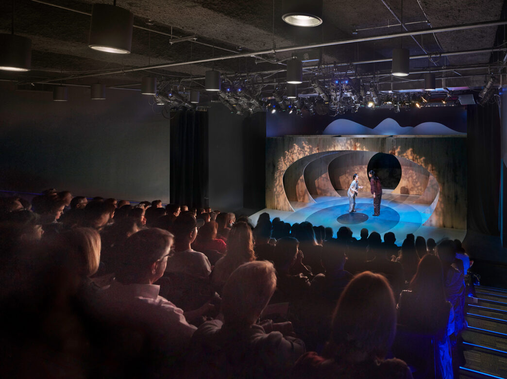 The audience sits in theater seats while facing a performance on stage at The Drake.
