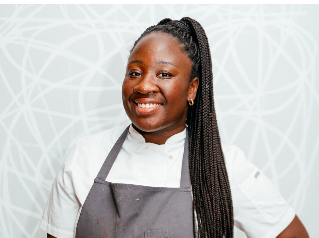 Chef Nana Araba Wilmot poses for a headshot while smiles at the camera. She wears a gray apron over a white chef's shirt.