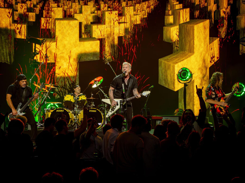 The band Metallica performs on stage in front of a crowd. The lead singer plays an electric guitar and sings into a microphone.