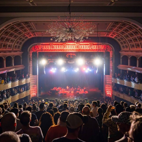 Una banda toca en el escenario del The Met en Filadelfia, mientras los asistentes al concierto miran hacia el escenario. Del techo cuelga una lámpara de araña y el escenario está iluminado con luces rojas.