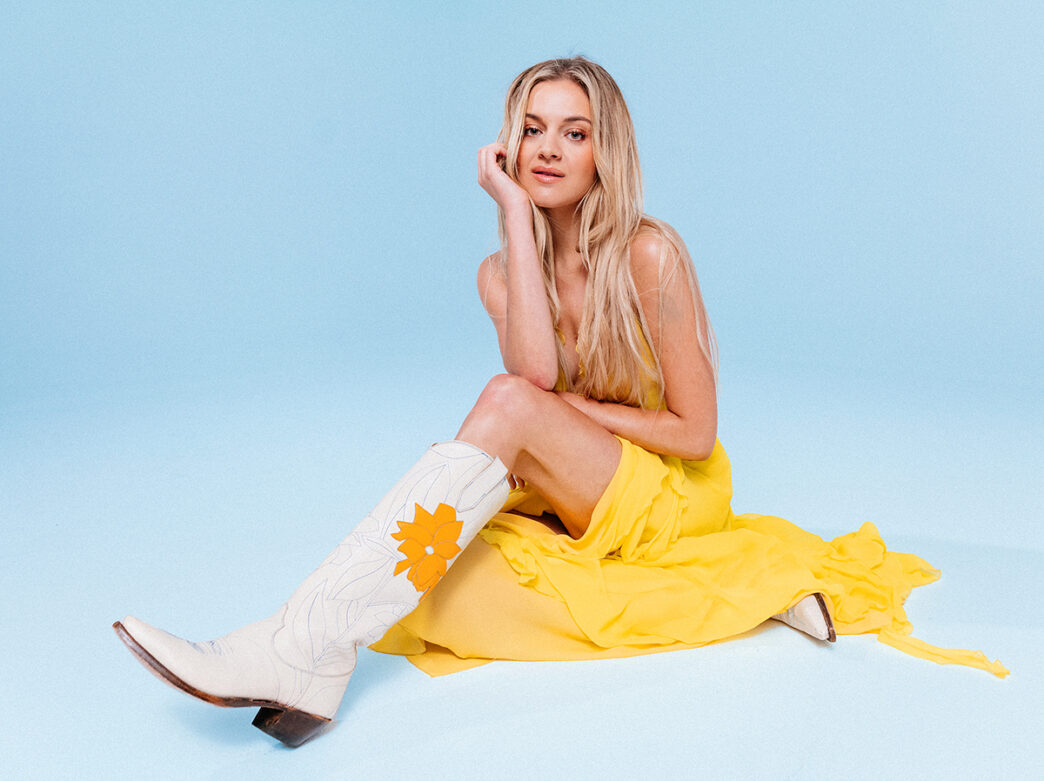 Singer-songwriter Kelsea Ballerini sits on the floor against a light blue backdrop while wearing a long yellow dress and a pair of white cowboy boots.