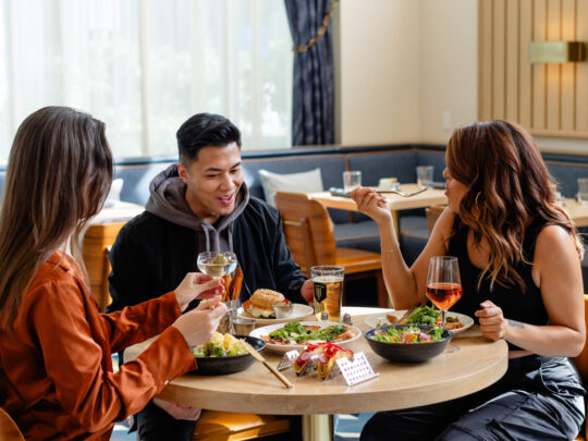 Three friends sit around a circular wooden table inside a stylish restaurant, enjoying salads, sandwiches and drinks.