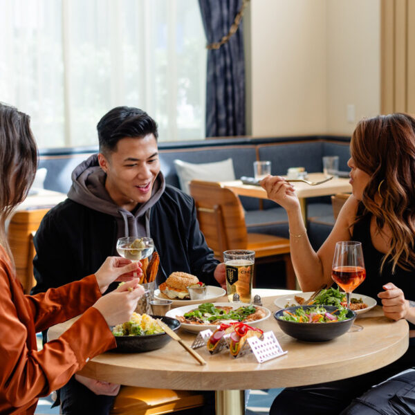 Three friends sit around a circular wooden table inside a stylish restaurant, enjoying salads, sandwiches and drinks.