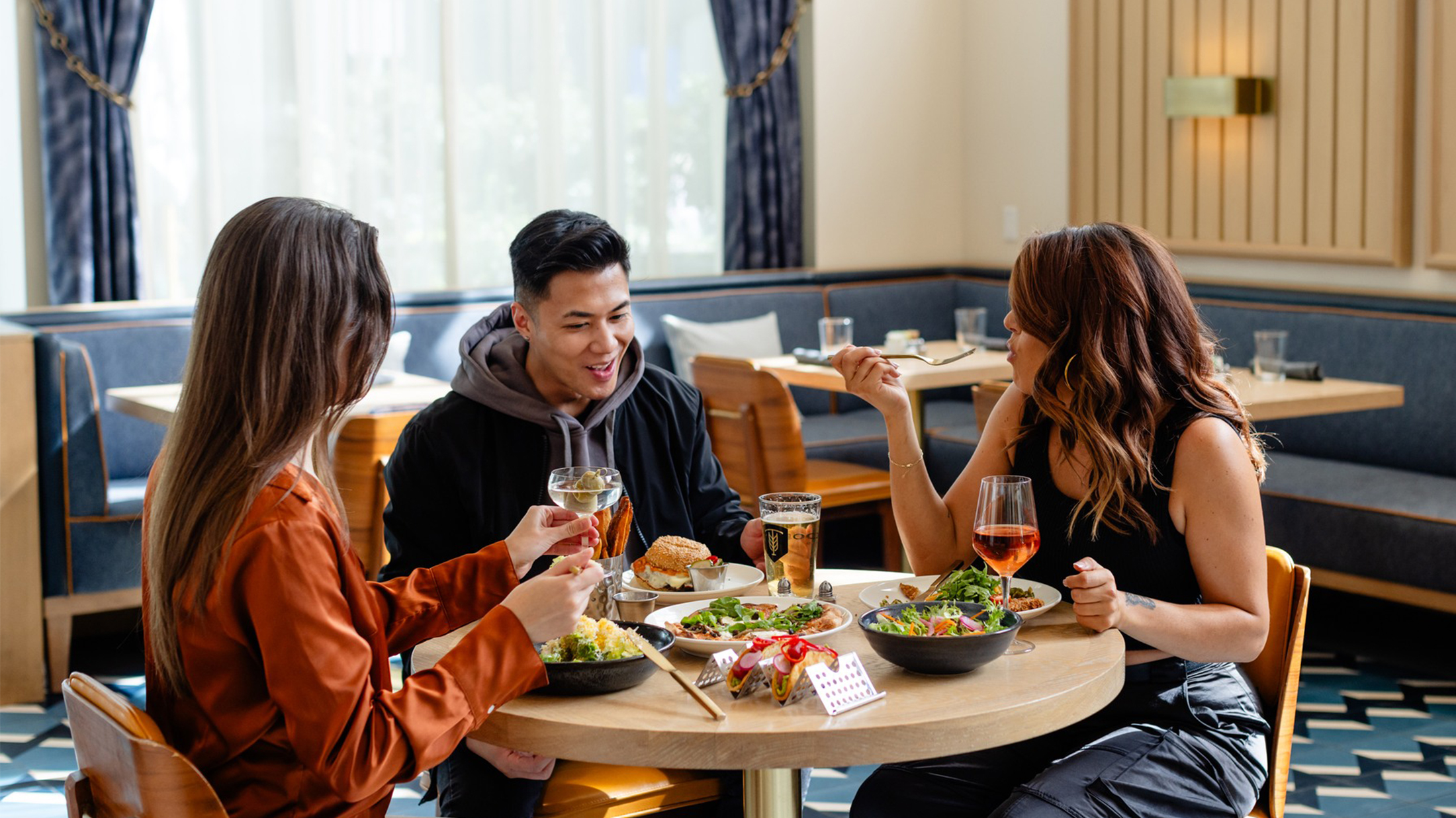 Three friends sit around a circular wooden table inside a stylish restaurant, enjoying salads, sandwiches and drinks.