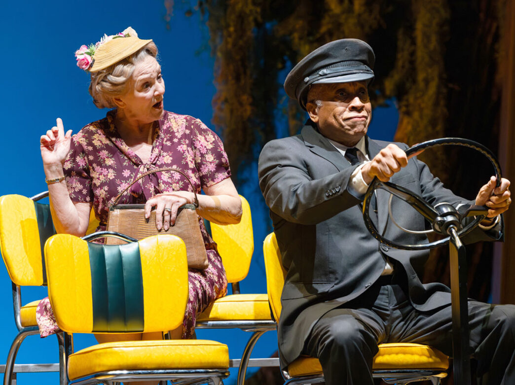An actress playing Miss Daisy and an actor playing Hoke perform on stage during a performance of Driving Miss Daisy. Miss Daisy sits in the back row of a vehicle while Hoke wears a black suit and drives.