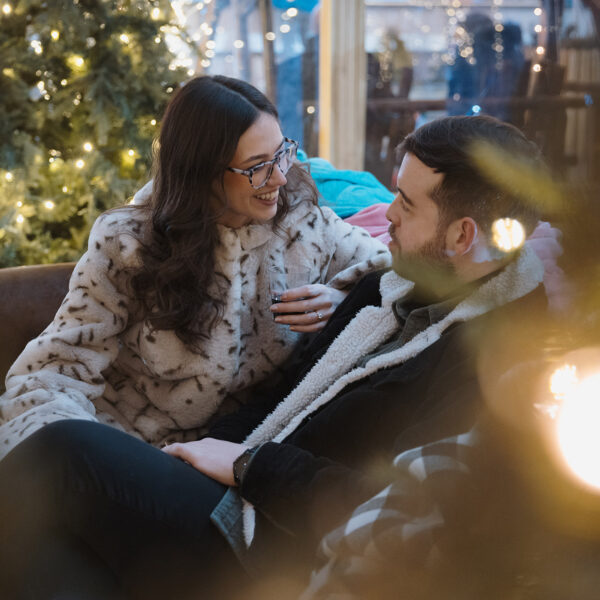 A cozy moment at Independence Blue Cross Riverrink Winterfest captures a couple seated together, smiling warmly at each other amidst twinkling holiday lights and festive greenery.