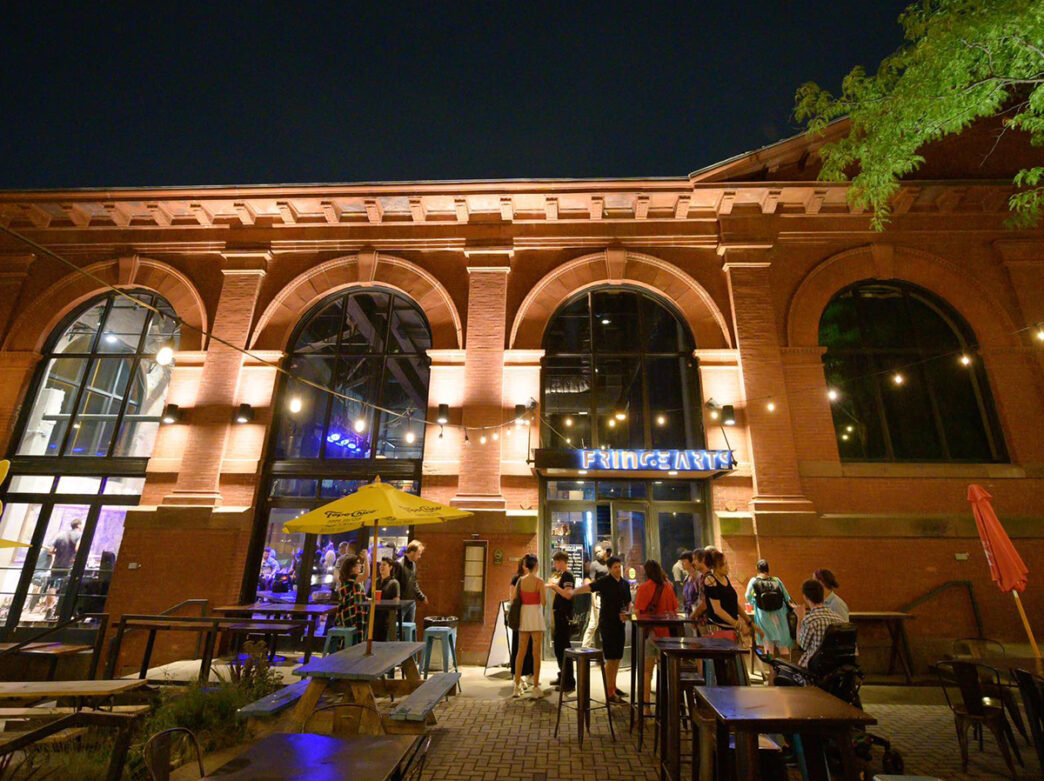 People stand outside of the FringeArts building at night. The red brick building has floor to ceiling windows and is illuminated by lighting.