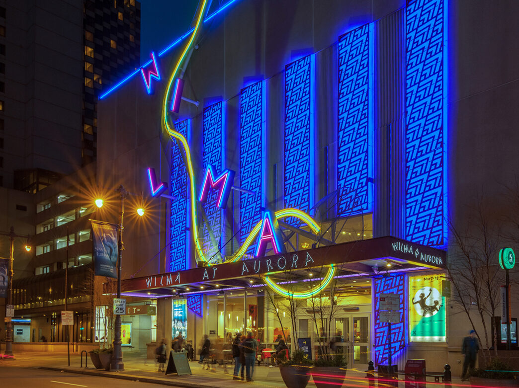 An exterior of shot of The Wilma Theater at night. The building is illuminated with blue lighting and a neon WILMA sign.