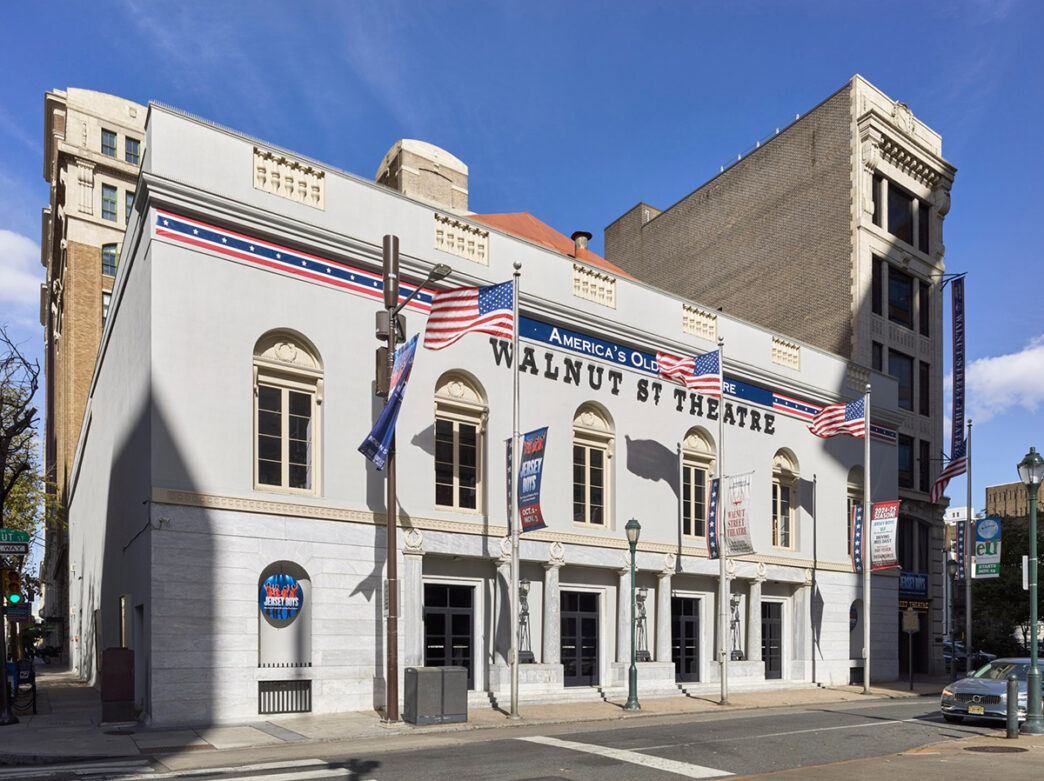 The exterior of Walnut Street Theatre. It is a light gray building with three American flags on flag poles.
