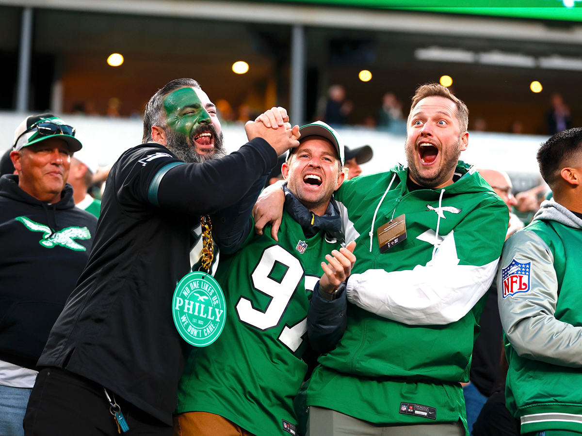 Three Philadelphia Eagles fans hug each other and celebrate while watching a game. One fan's face is painted white and green and the other two wear green Eagles attire.