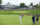 A golf swings at a golf ball on the fairway. Two caddies stand with the golfers bag. A crowd of people watching can be seen in the far distance.