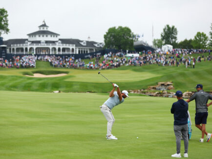 Un golfeur frappe une balle de golf sur le fairway. Deux caddies se tiennent à côté du sac du golfeur. Au loin, on aperçoit une foule de spectateurs.