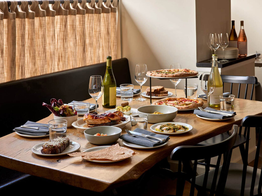 Various menu items are displayed on a rectangular wooden table at High Street in Philadelphia. Included are two pizzas, a half roast chicken, bread, two bottles of wine and more.