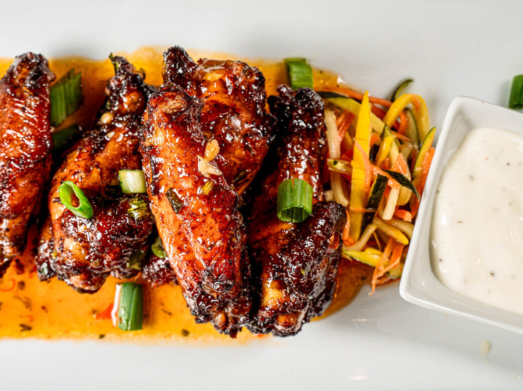 An order of chicken wings are served on a white plate with a size of pineapple-ranch dip and vegetable slaw. The chicken wings are smothered in sauce.