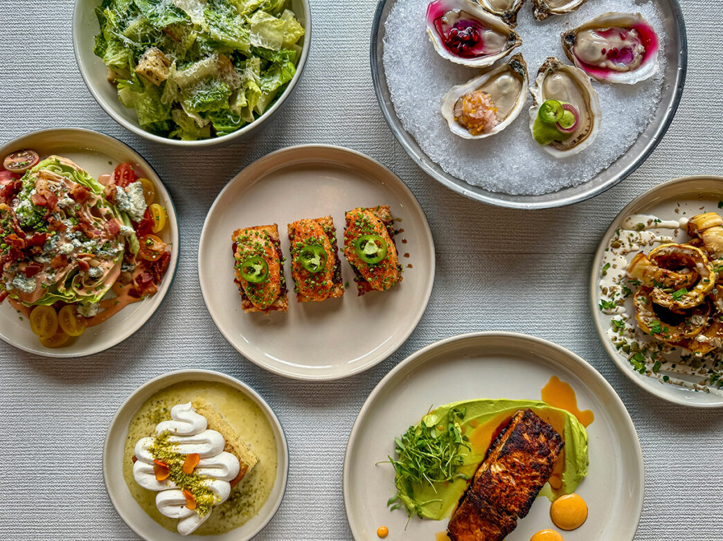 Menu items from Jules@Market are diplayed on a table with a white tablecloth. Items include salmon crispy rice, a filet blacked red drum, Caesar salad, pistachio tres leches, oysters and more.