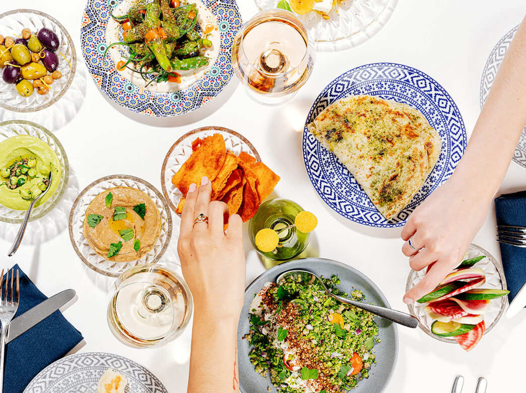 An overhead shot of a table with various menu items displayed. Hands reach toward the food items. Hummus, olives, naan and salads are on the table.