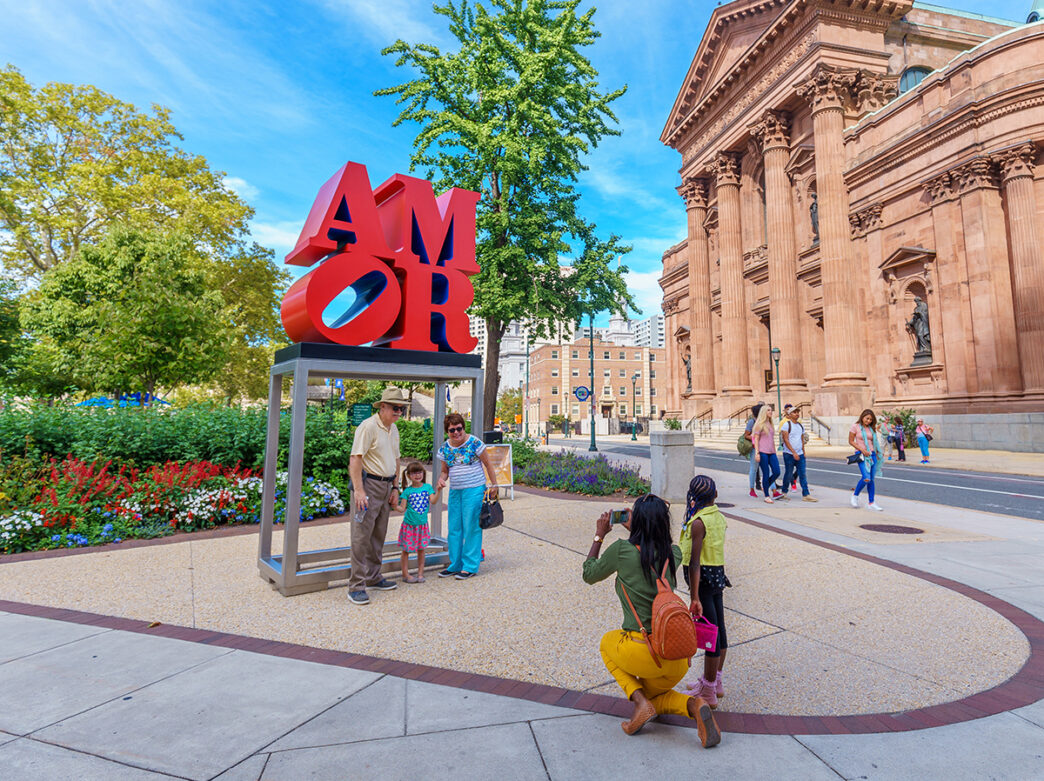 A child stands between two adults while posing for a photo in front of the AMOR Statue in Philadelphia. A person kneels on the ground to take their photo with a phone. Greenery and buildings are in the background.