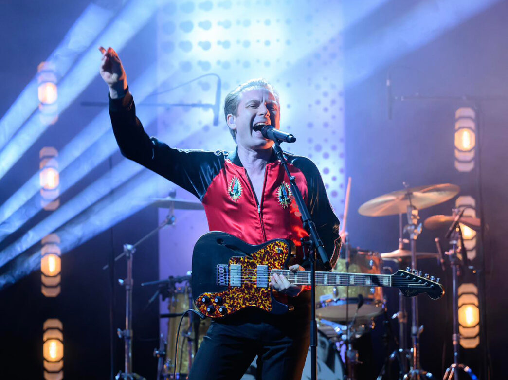 Lead singer, Alex Kapranos, of indie rock band Franz Ferdinand sings into a microphone while playing an electric guitar during a concert. A band member can be seen in the background playing the drums.