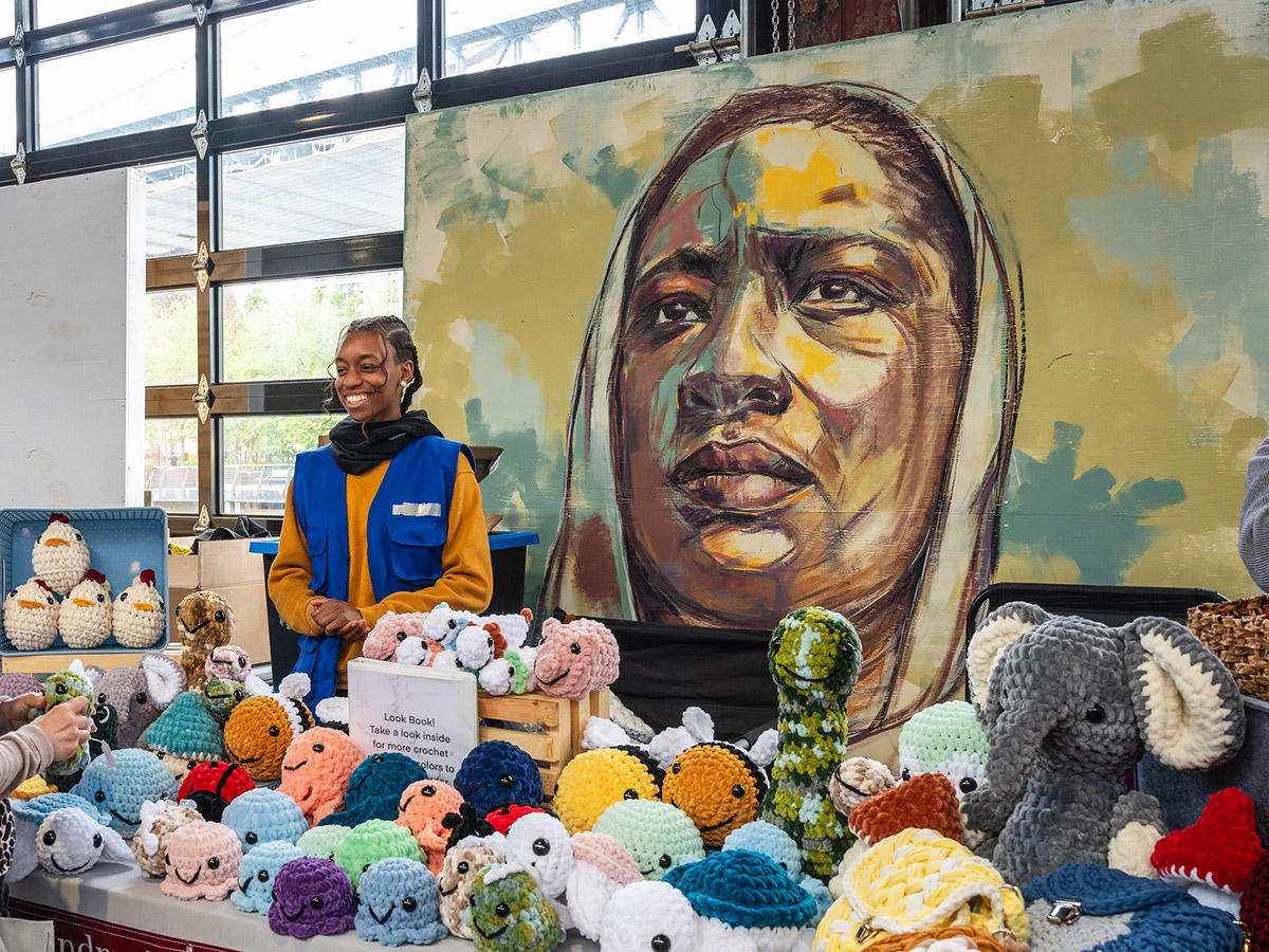 Een verkoper gekleed in een oranje sweater en blauw vest staat achter haar tafel met schattige en kleurrijke gehaakte pluchen. Achter haar is een bruin met blauwe muurschildering van een vrouw te zien.