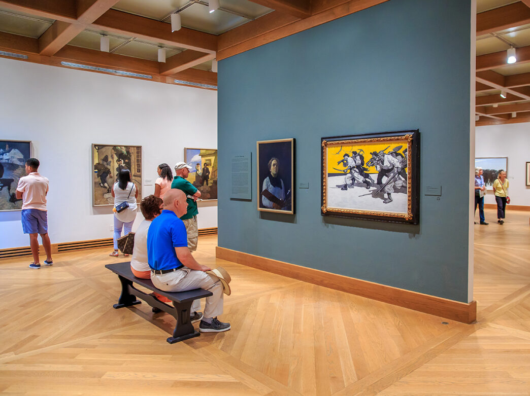 Two people sit on a bench and look at two paintings hung on a gallery wall at the Brandywine Museum of Art. Other people walk around and look at the artwork.