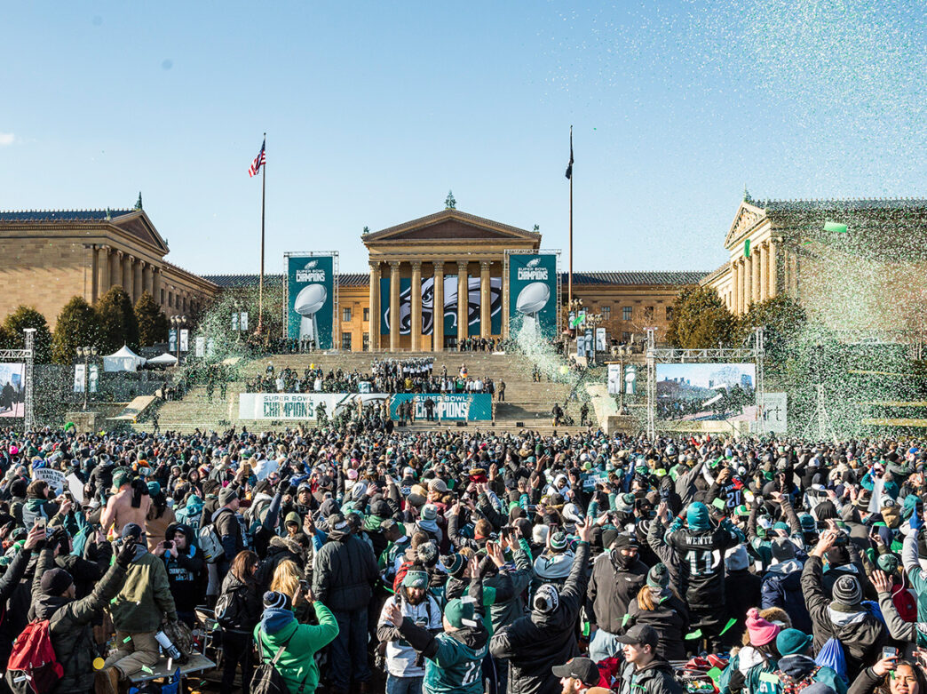 A crowd of stand at the base of the steps of the Philadelphia Museum of Art during the Super Bowl Celebration in Philadelphia. Banners of the Vince Lombardi Trophy and Philadelphia Eagles logo hang from the Art Museum's exterior.