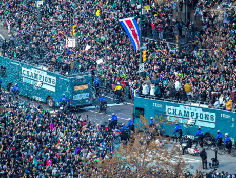 Two open open-air buses driving along the Eagles Super Bowl Parade route while crowds of people cheer from the sidewalks. Confetti is throw into the air.