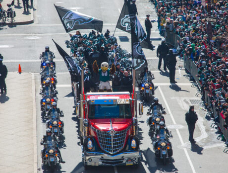 Flanked by police motorcycles, a red truck is adorned with Eagles flags and carries the team's mascot Swoop, cheerleaders and staff down the street as a crowd of fans watches.