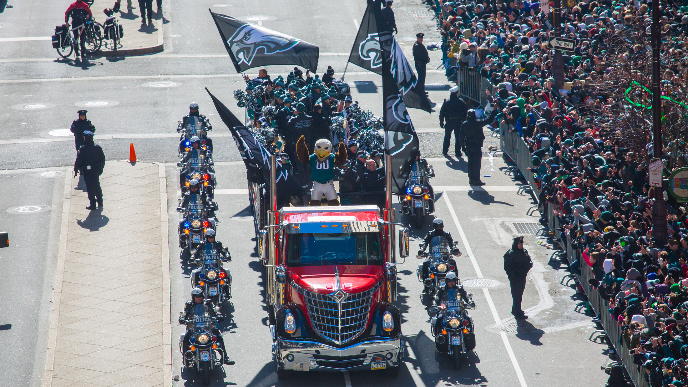 Flanked by police motorcycles, a red truck is adorned with Eagles flags and carries the team's mascot Swoop, cheerleaders and staff down the street as a crowd of fans watches.