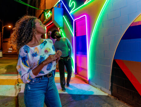 A group of friends explores the Electric Street Mural, an alleyway illuminated by vibrant neon lights arranged in geometric patterns on a colorful painted wall, at night.