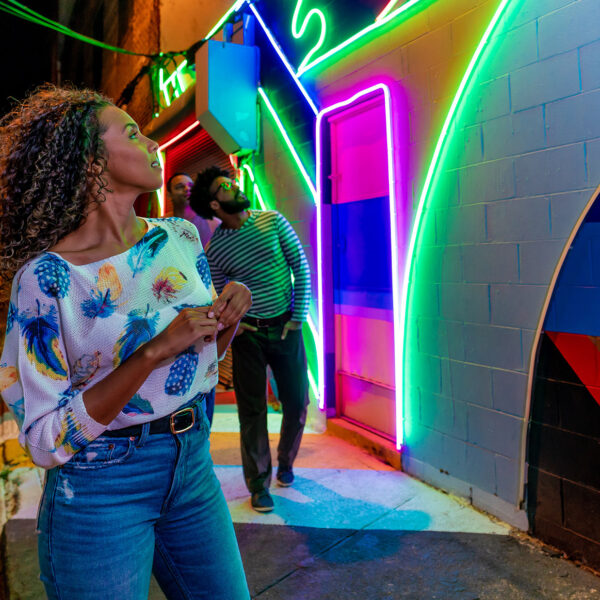 A group of friends explores the Electric Street Mural, an alleyway illuminated by vibrant neon lights arranged in geometric patterns on a colorful painted wall, at night.